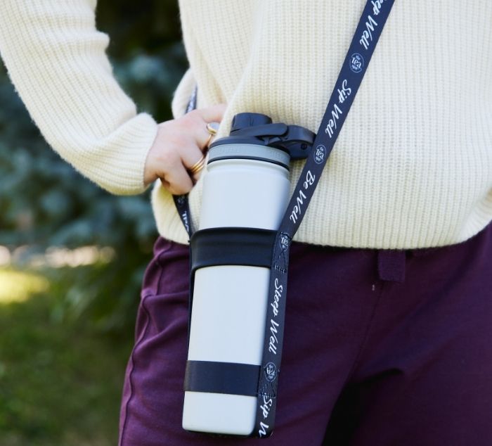 A white insulated tea travel bottle with a black lid and black silicone sleeve, carried on a person's hip by a black bottle strap printed with the words 'Steep Well'.
