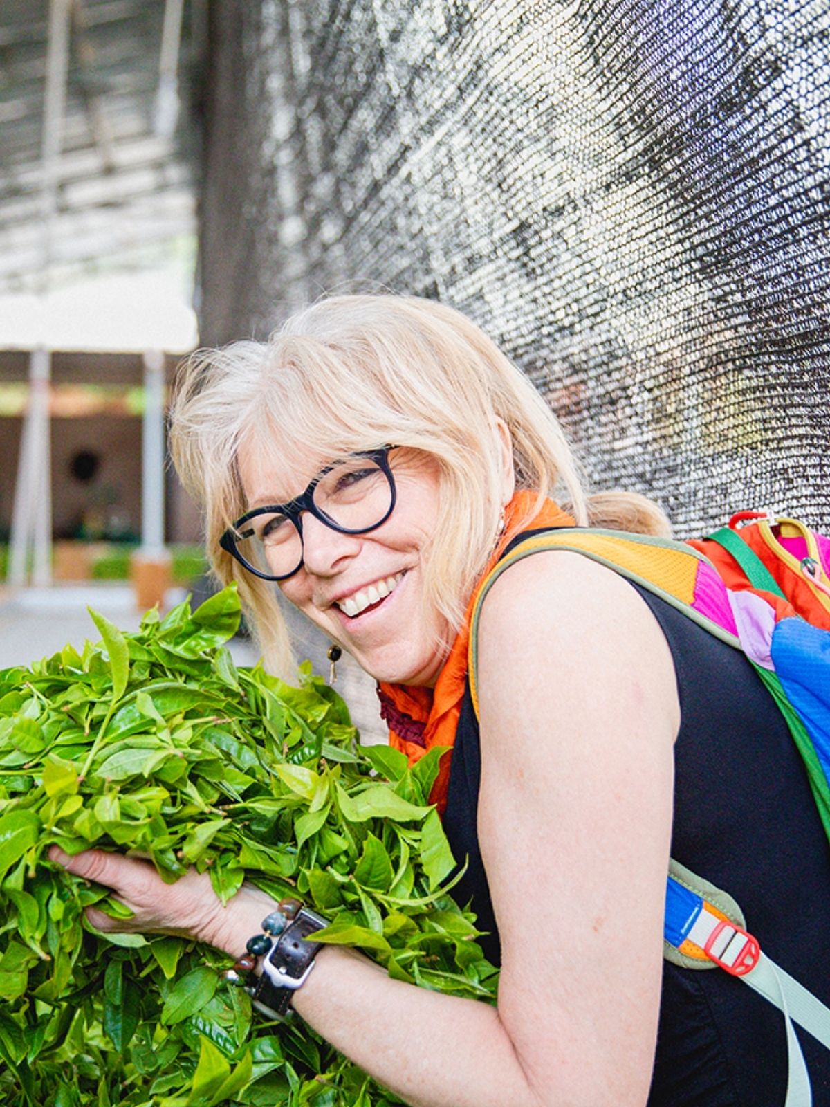 Maria Uspenski leans over a large bunch of fresh green tea leaves, wearing a black sleeveless top, an orange scarf, and a colorful backpack against a textured metallic background.
