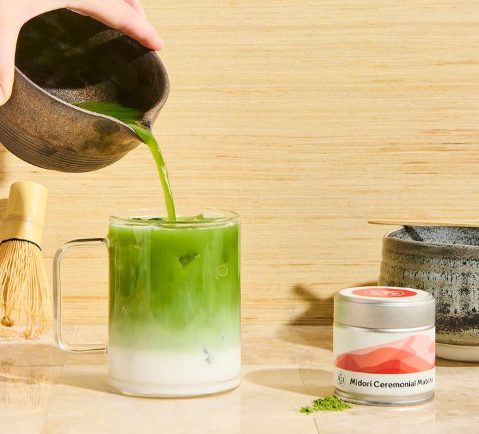 a hand pours Midori Matcha green tea into mug to make a latte next to tea tin, matcha bowl and whisk