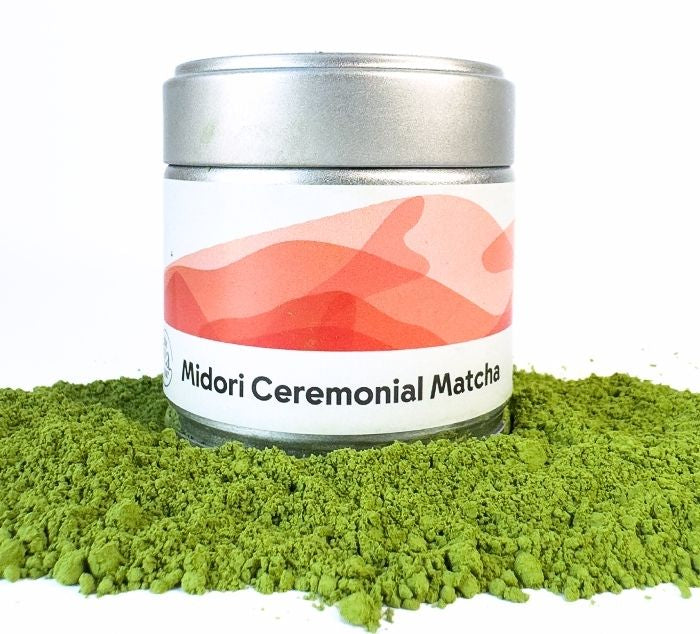 A silver metal tin of Midori Ceremonial Matcha green tea powder, surrounded by a pile of vibrant green matcha powder on a white background.