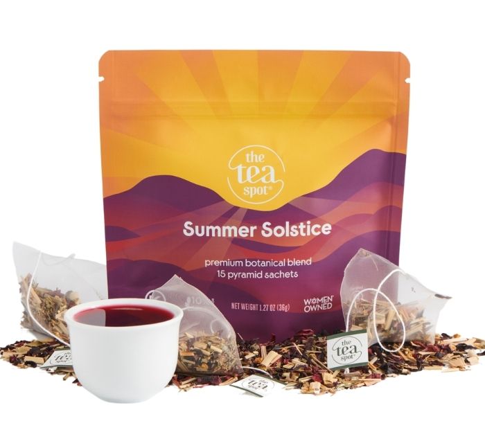 The Tea Spot Summer Solstice herbal tea in pyramid sachets with a red cup of brewed tea and whole botanical ingredients on display.