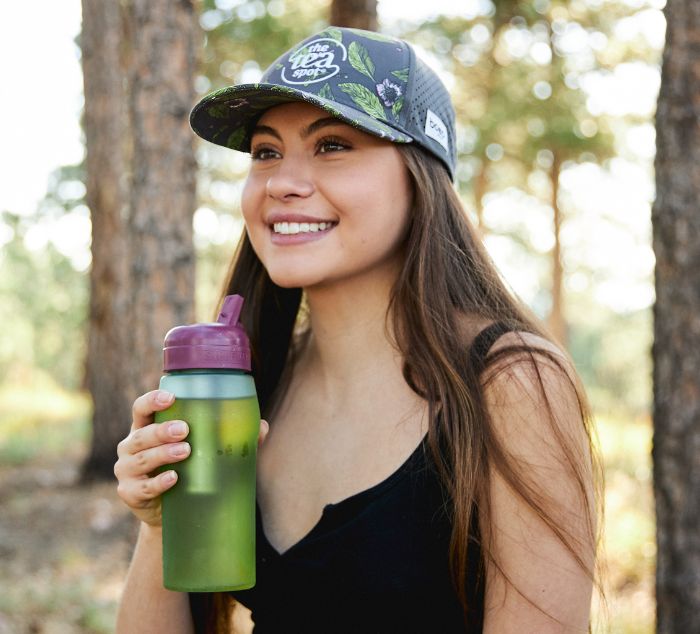 A woman outdoors in a forest setting, smiling and holding a green reusable water bottle with a purple lid. She is wearing a dark patterned baseball cap with the tea spot logo on it, and a black tank top.